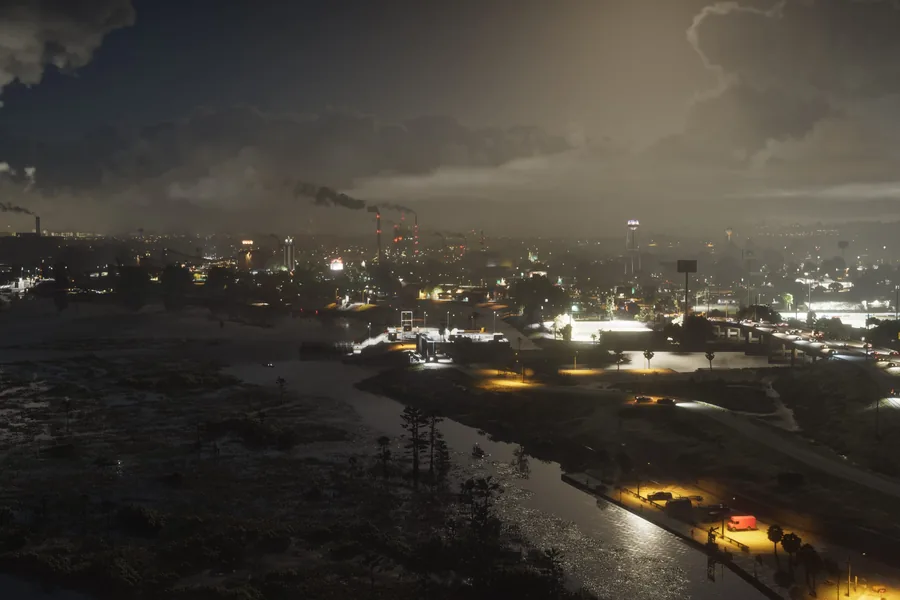 Ambrosia at night seen from above: a lit industrial town with smoking chimneys and a water tower, a highway on the right and dark marshland in the foreground.