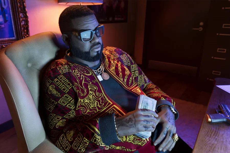 A bearded man in glasses, wearing a burgundy-and-gold baroque jacket and a gold chain, sits in an office armchair holding a wad of banknotes in a purple-lit room.