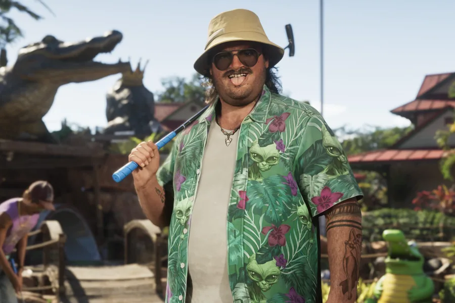 A heavyset man in a beige bucket hat, sunglasses and a green Hawaiian shirt patterned with cats, holding a putter on a minigolf course decorated with an alligator statue.