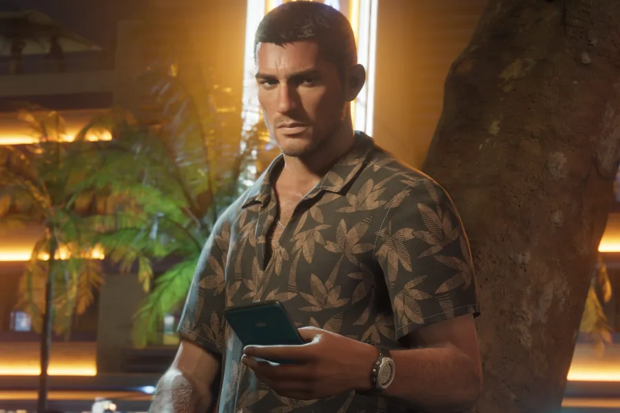 Jason Duval leans against a tree on a neon-lit avenue at night, a phone in his hand, wearing a leaf-patterned short-sleeved shirt.