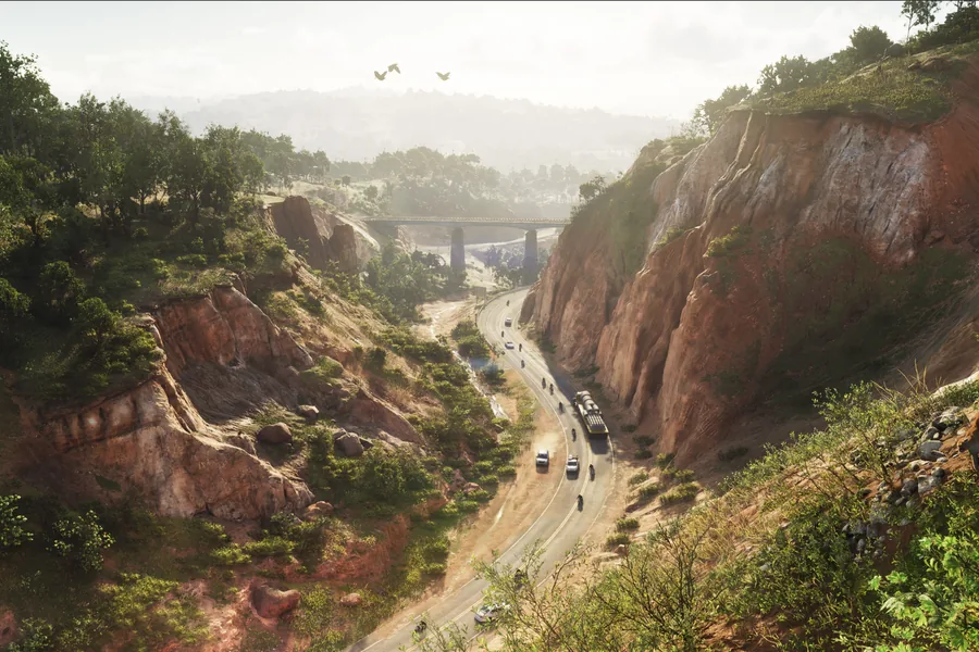 A mountain road winding along the floor of a red-earth gorge, a few vehicles in single file, a viaduct spanning the valley in the distance and wooded hills in low, raking light.