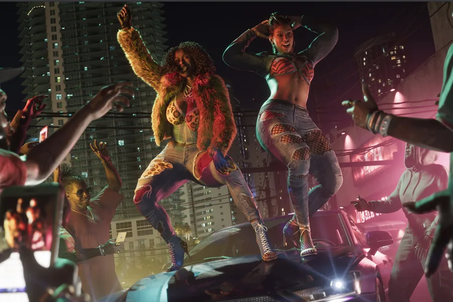 Two women dance standing on a car roof at night, surrounded by a crowd filming them on phones, under pink neon and residential towers.